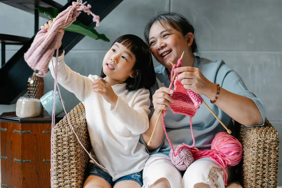An Asian grandmother laughing as she knits with her granddaughter